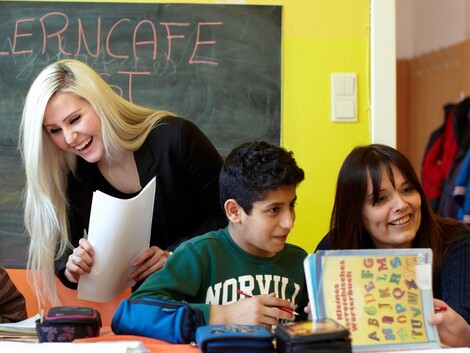 Schulkinder sitzen an einem Tisch und machen Aufgabe, daneben steht eine junge Frau mit Zetteln in der Hand.