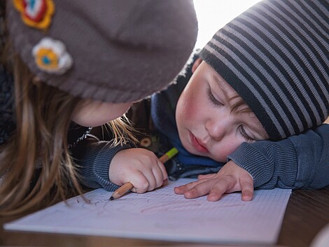 Zwei Kinder sitzen an einem Tisch, eines der beiden Kinder hat einen Stift in der Hand und sein Kopf liegt auf seiner Hand, sein Blick ist traurig zu Boden gerichtet. Beide tragen eine Mütze.